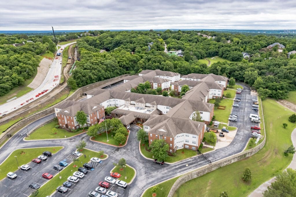 DJI_0152-11207123 - Grace Management Aerial view of an upscale senior living community surrounded by lush greenery and ample parking spaces.
