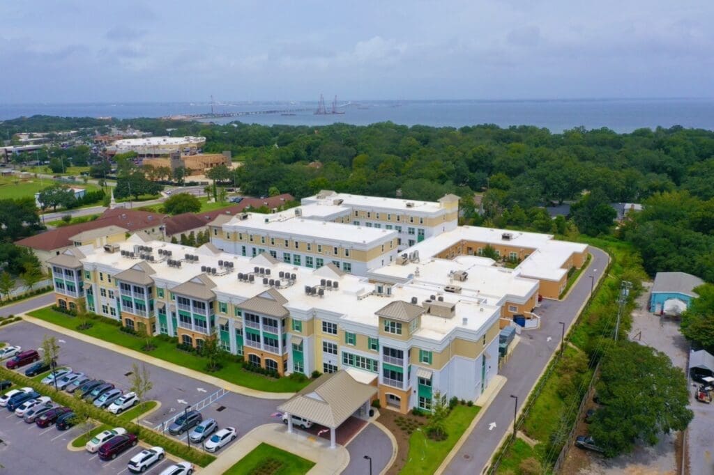 DJI_0620-Large - Grace Management Aerial view of a multi-story beige apartment complex with green accents, surrounding parking lots, and manicured landscaping near a wooded area and distant shoreline.