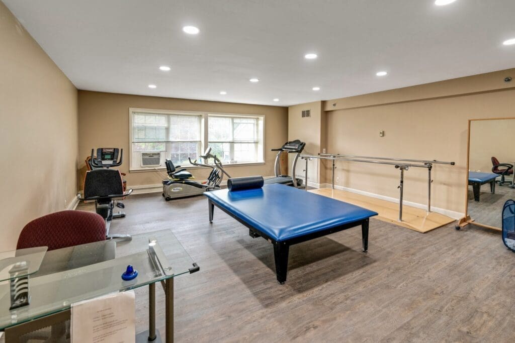 Spacious therapy room featuring a blue treatment table, exercise equipment, and large windows for natural light.