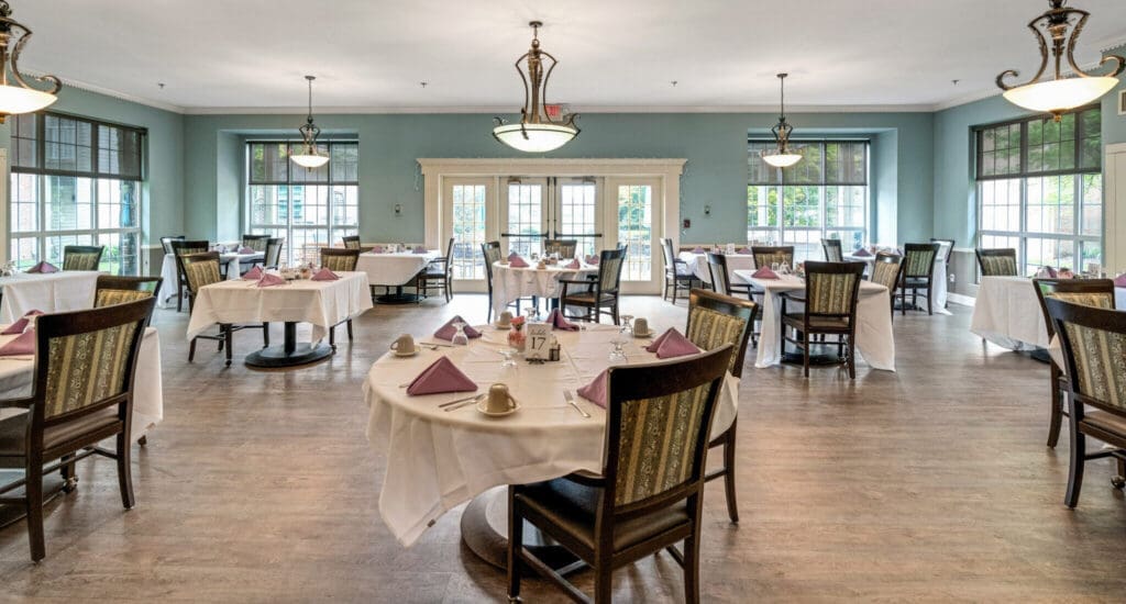 Elegant dining room featuring neatly set tables with white linens and lavender napkins, illuminated by stylish chandeliers.