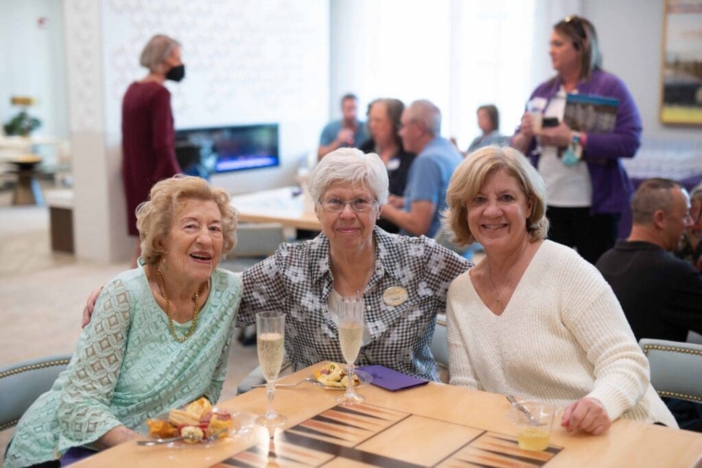 Three smiling women enjoying refreshments together in a vibrant senior living community gathering.
