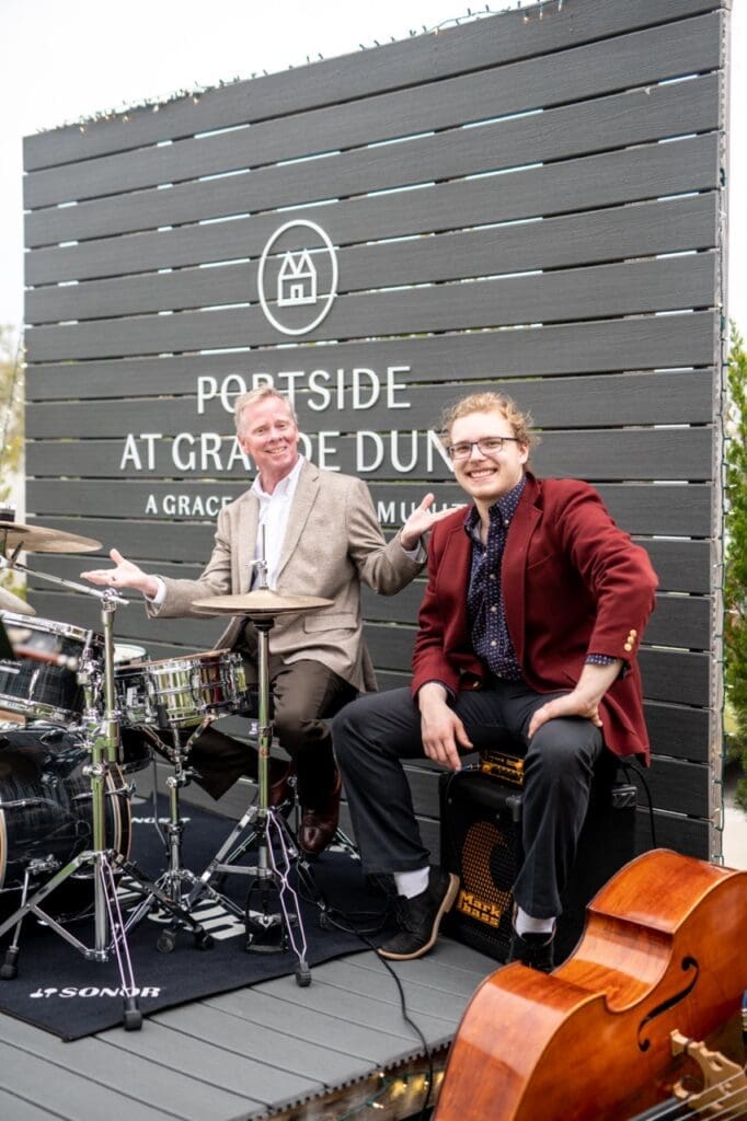DSC1709-Large - Grace Management Two musicians joyfully playing instruments in front of a stylish sign for Portside at Grande Dunes, showcasing vibrant community life.