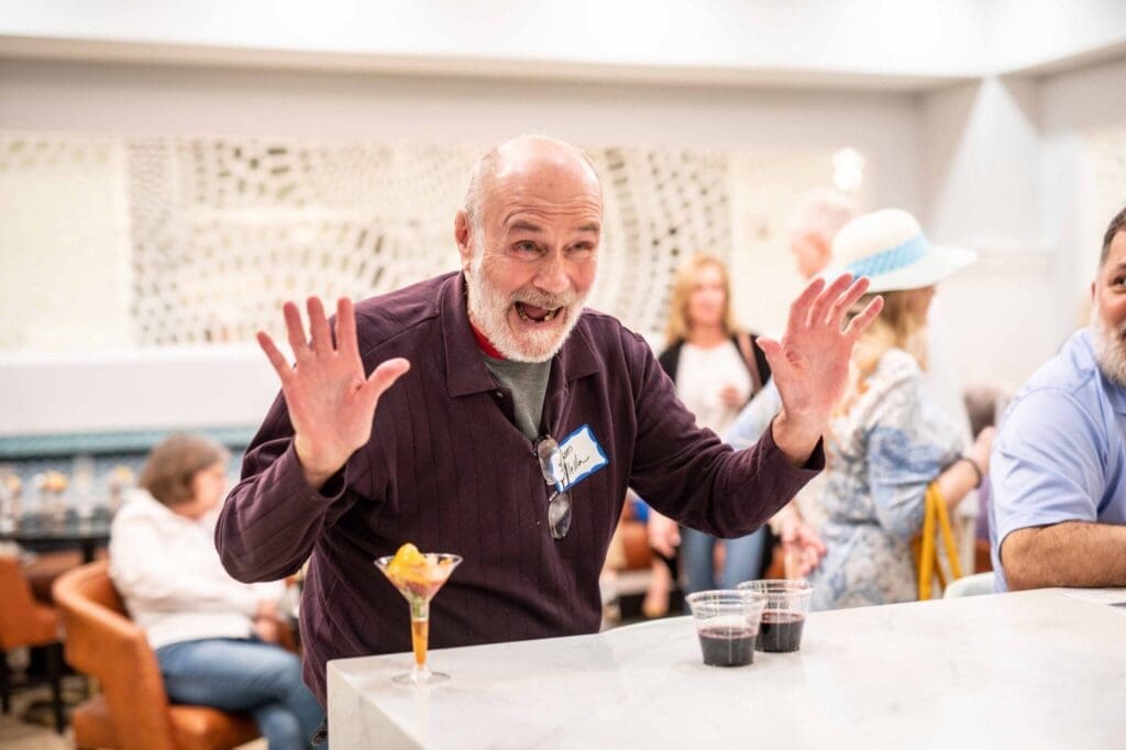 Joyful senior man enthusiastically gestures while enjoying a vibrant social event, with cocktails on the bar in a stylish setting.