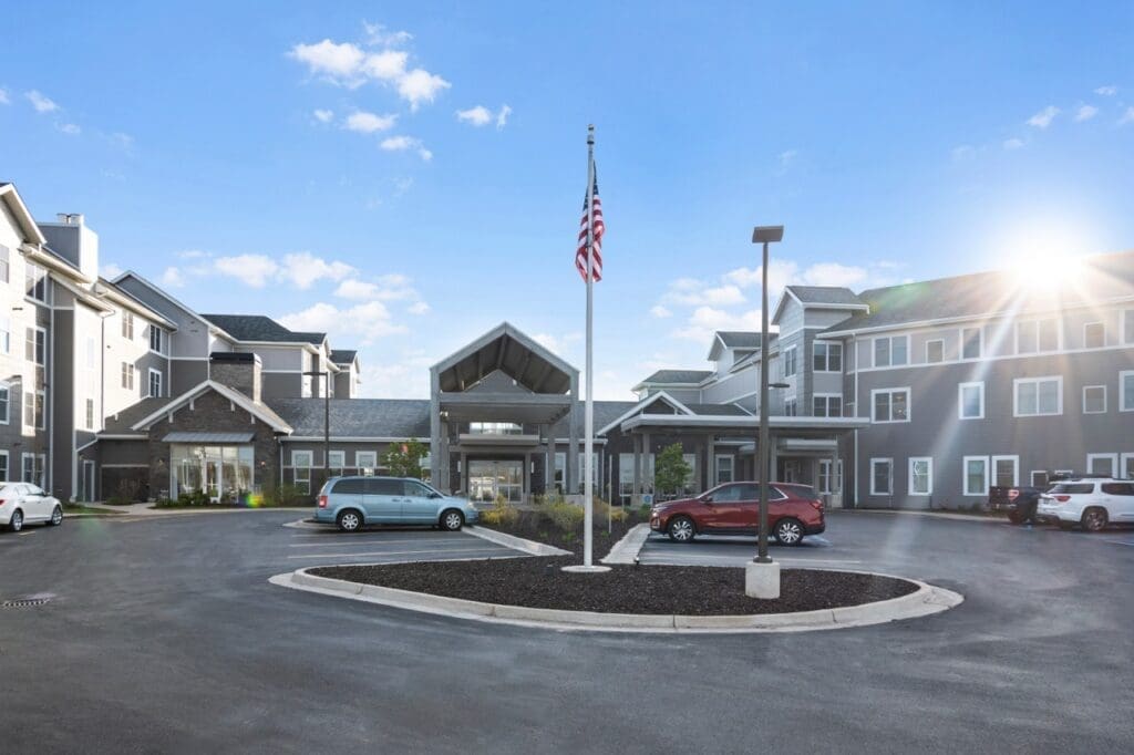 DSC4311-Large - Grace Management Modern senior living community entrance with vibrant landscaping, ample parking, and an American flag against a bright sky.