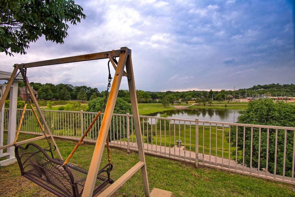 E-TownVillage_TulsaOK_BrownSwing2 - Grace Management Scenic view from a swing overlooking a serene pond, lush greenery, and vibrant landscape under a partly cloudy sky.