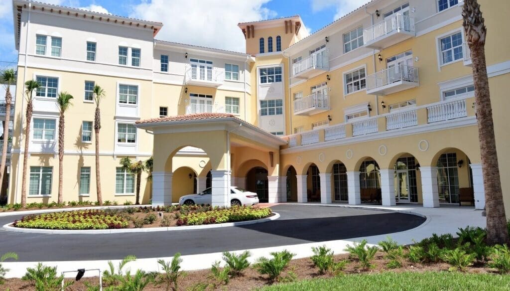 Exterior-Front-Large-e1750256703159 - Grace Management Elegant senior living community entrance with landscaped gardens and a welcoming porte-cochère under bright blue skies.