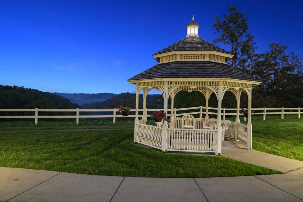Charming white gazebo in a lush, landscaped setting, with vibrant flowers and serene mountain views under a twilight sky.