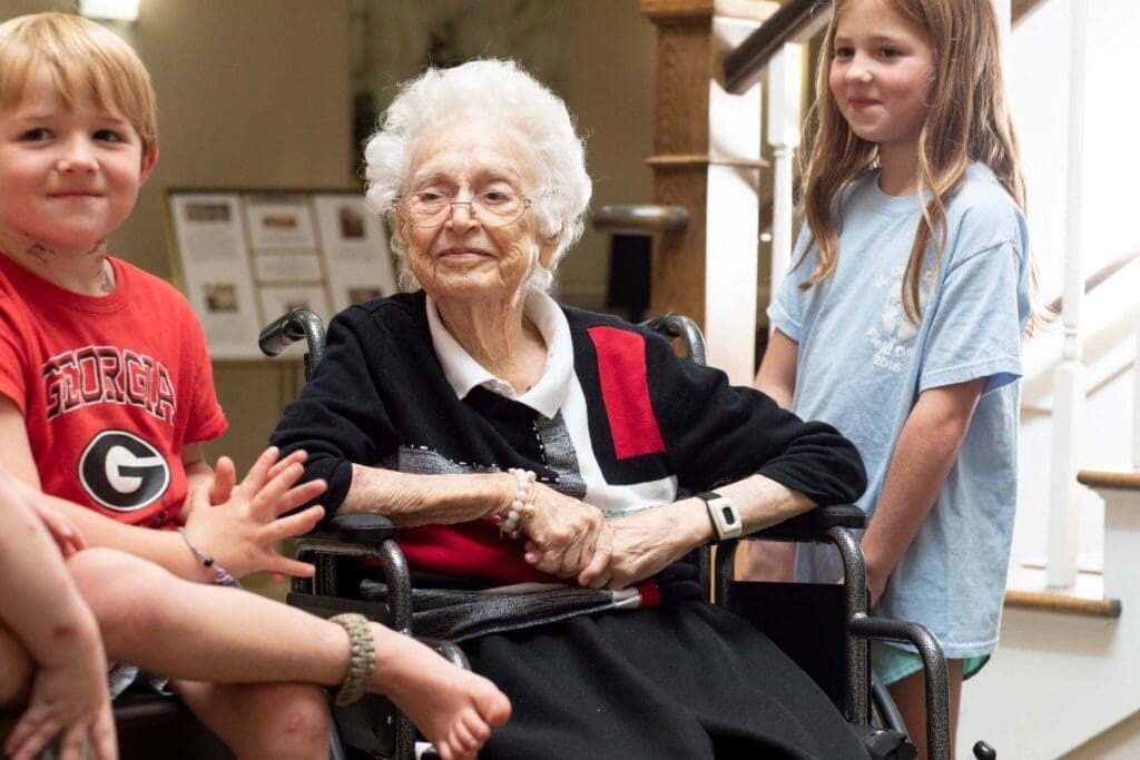 An elderly woman in a wheelchair smiles warmly as two children interact with her in a welcoming, lively setting.