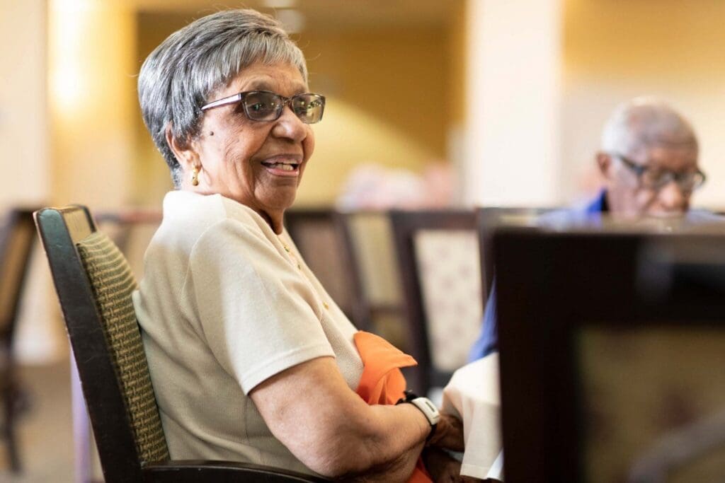 Joyful senior woman with glasses smiles warmly while seated in an inviting dining area, enjoying community moments.