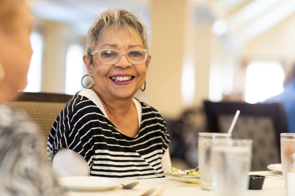 Smiling senior woman with glasses in a dining setting, enjoying a meal with a vibrant atmosphere and compassionate company.