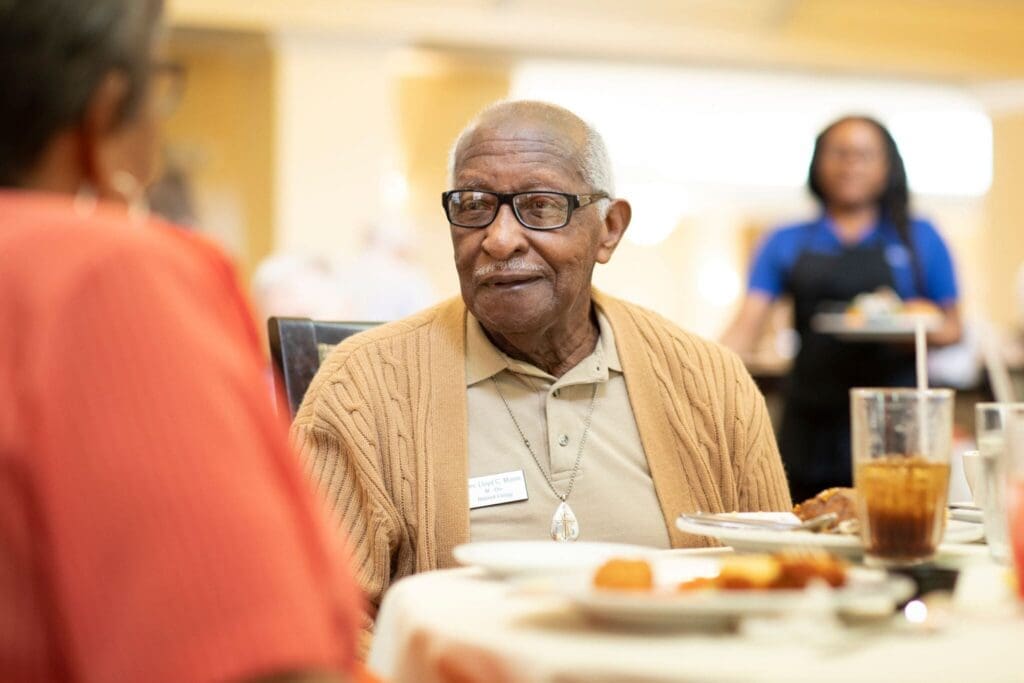 A senior gentleman with glasses enjoys a meal, engaging warmly with a companion in an inviting dining setting.