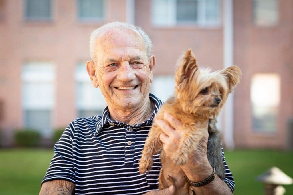 Grace_Management_Memphis_2018-331-2048×1365 - Grace Management Smiling elderly man in a striped shirt holds a small brown dog outdoors with a brick building in the background.