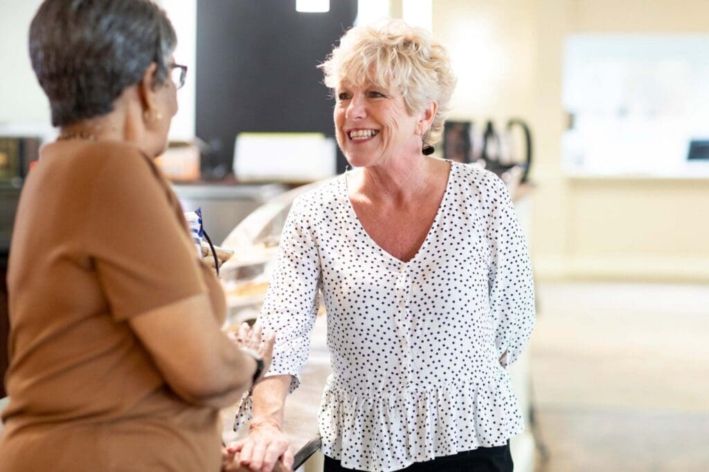 Two smiling women engage in cheerful conversation at a stylish café, surrounded by a warm, inviting atmosphere.