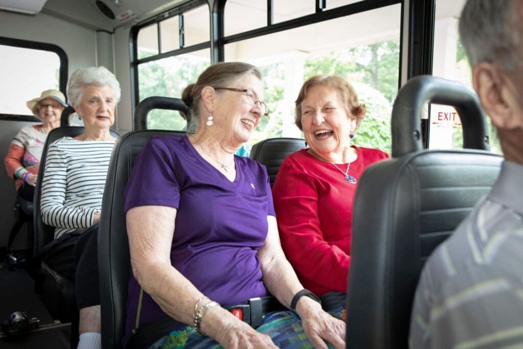 Two smiling senior women share a joyful moment on a bus, surrounded by fellow residents enjoying their vibrant outing.