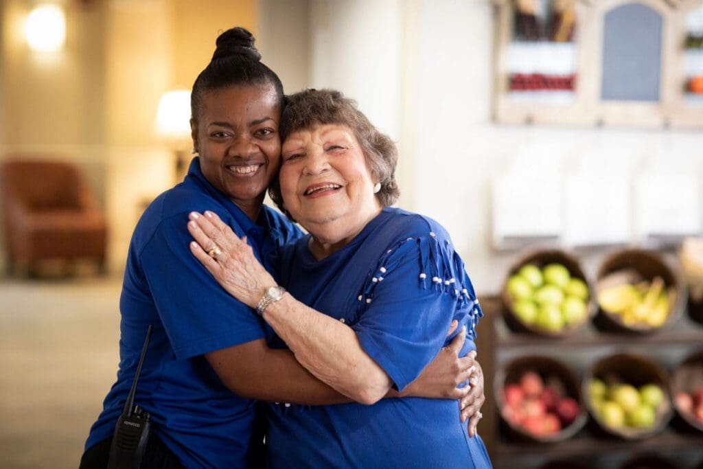 A staff member joyfully embraces a smiling senior resident in a vibrant common area with fresh fruit displays.