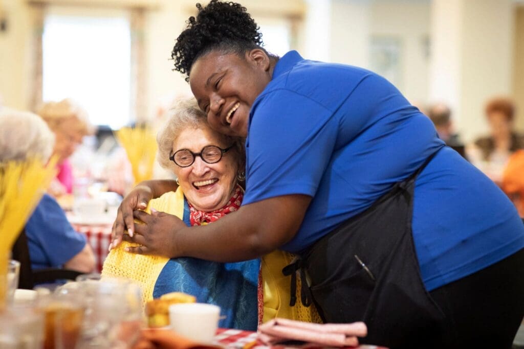 A caregiver joyfully embraces a smiling senior woman in a vibrant dining setting filled with happy residents.