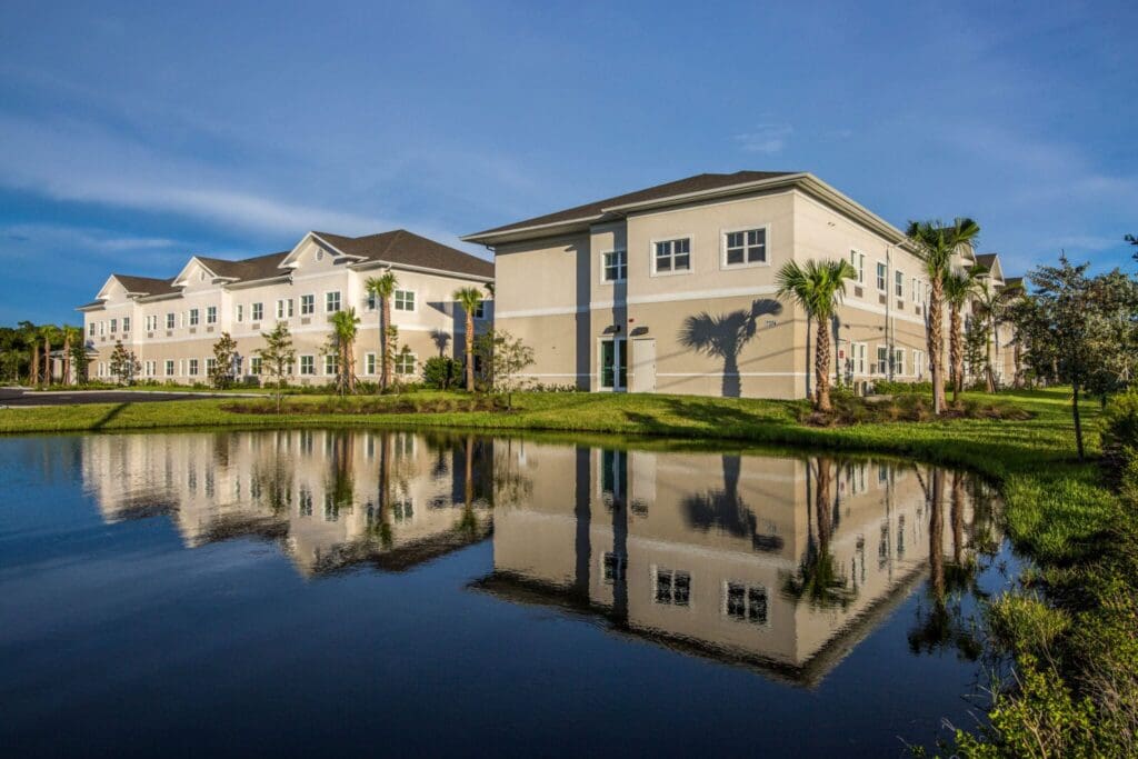 Heritage-Oaks1805-2048x1366 - Grace Management Modern senior living residence with vibrant landscaping, palm trees, and serene water reflection under a clear blue sky.