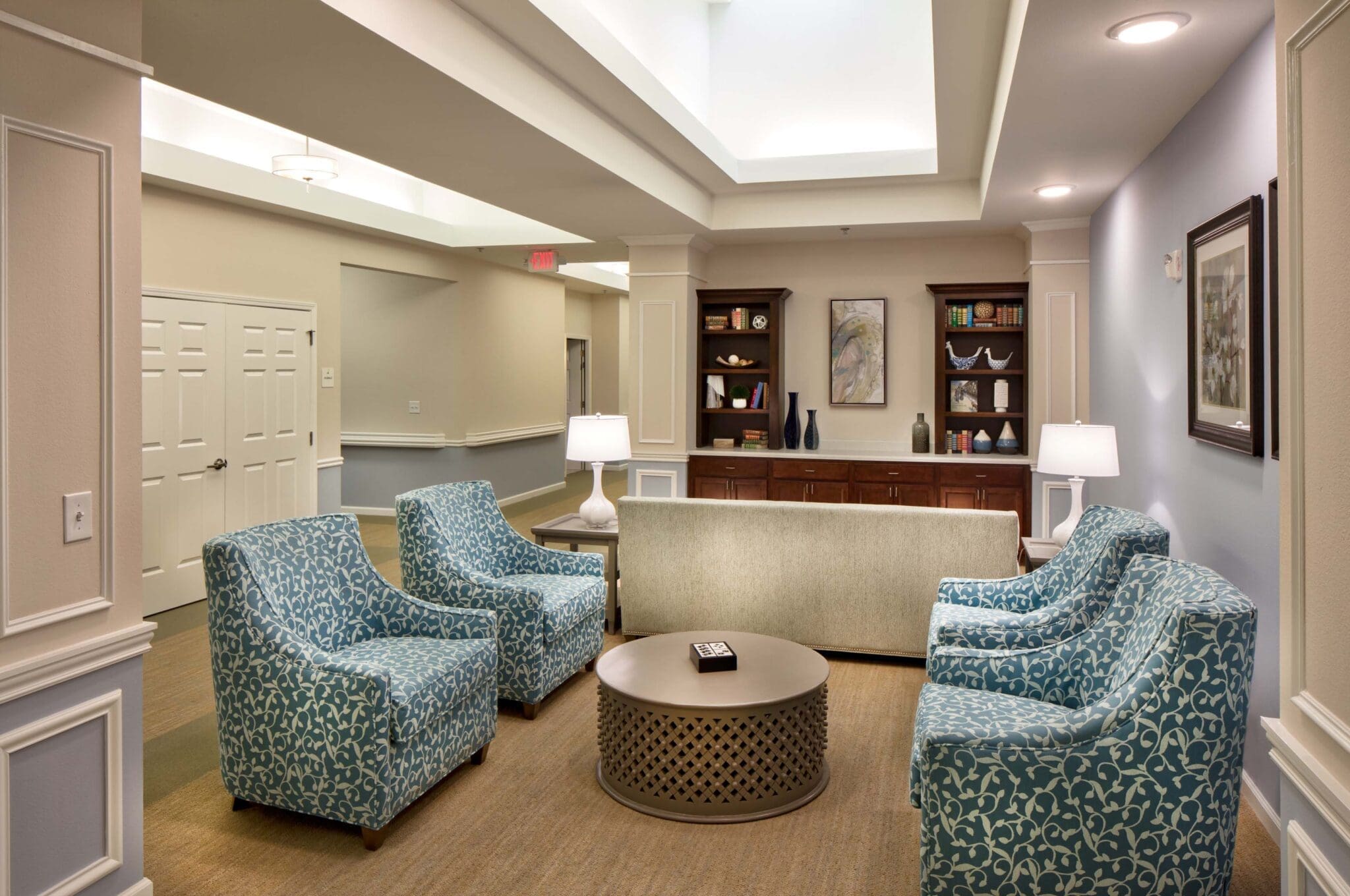 A sitting are with four blue sofa chairs, a circular coffee table between them, and a bookshelf to their right, at Twin Rivers, an assisted living and memory care community near Garland, TX.