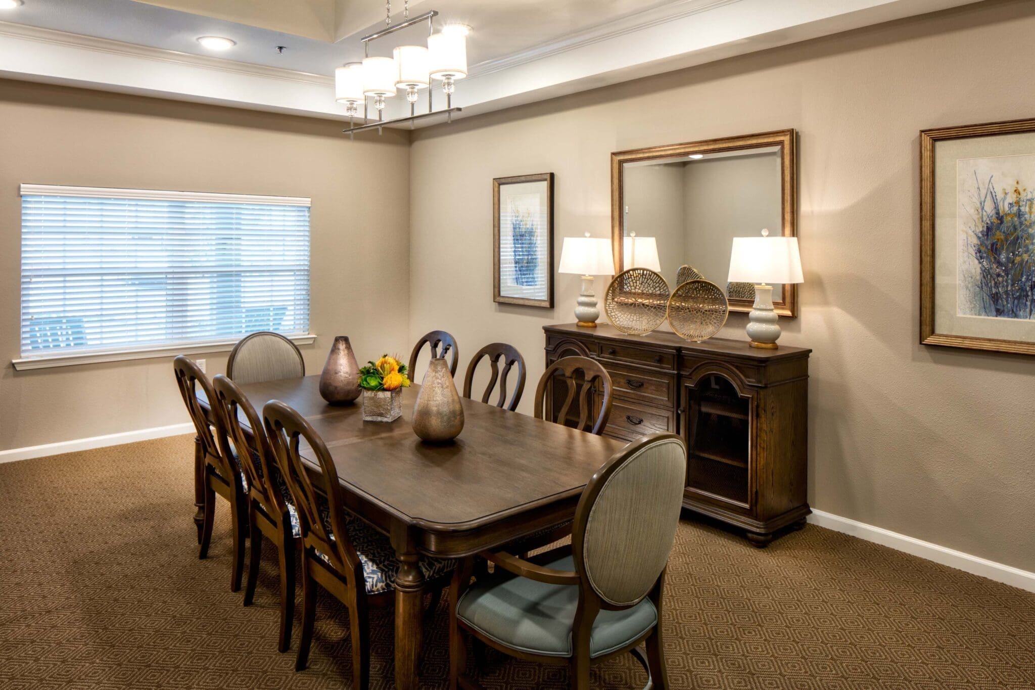 Lifestyle Options - Grace Management A long, dark-brown dining table with eight chairs, two table lamps, and a small flower bouquet on the table, a dresser to the right, underneath a mirror, and two lit-up lamps on the dresser at Twin Rivers, an assisted living and memory care community near Plano, TX.