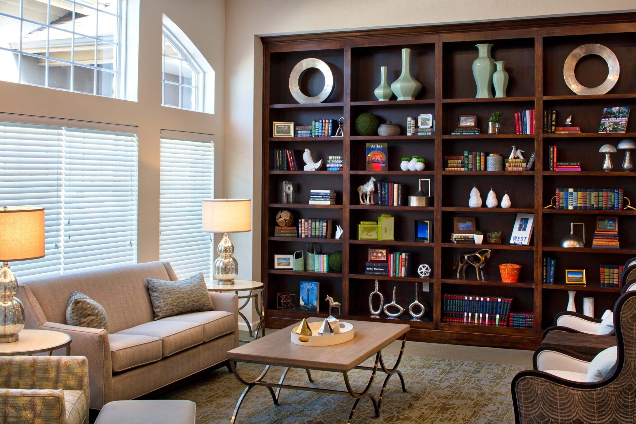 Events - Grace Management A sitting area with a beige couch and two sofa chairs placed in front of the couch, with the coffee table in between them, and a bookshelf full of vases, books, and other things to the right, against the wall at Twin Rivers, an assisted living and memory care community near Plano, TX.