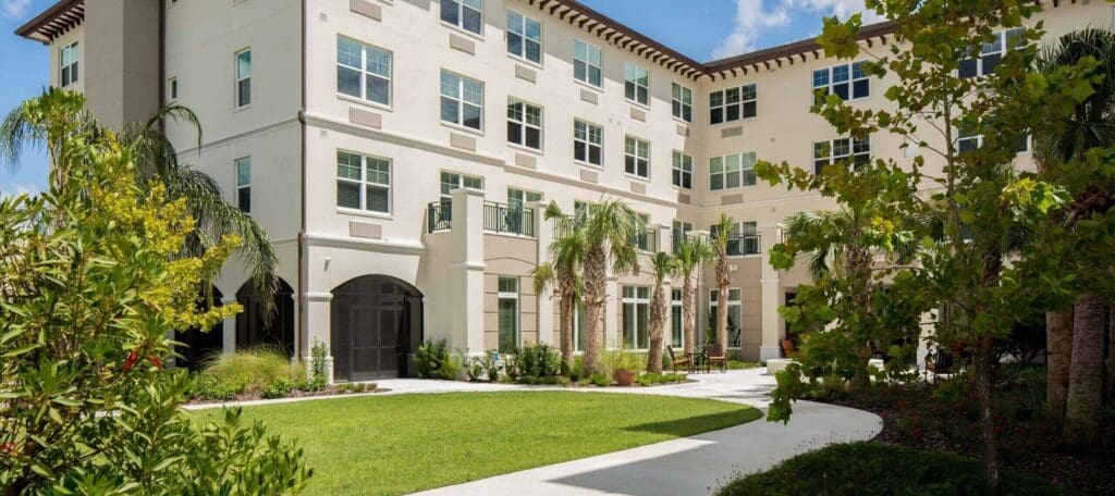 Elegant senior living community featuring lush gardens, palm trees, and modern architecture under a bright blue sky.