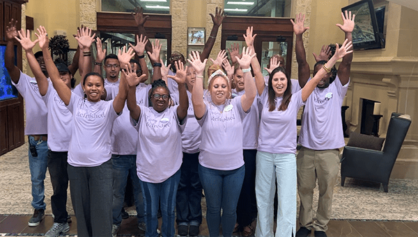 Team members in purple shirts celebrate joyfully, raising their hands in a welcoming senior living community lobby.