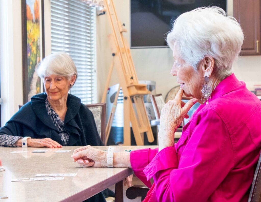Two elegantly dressed senior women engage in a card game, enjoying a vibrant atmosphere filled with laughter and connection.