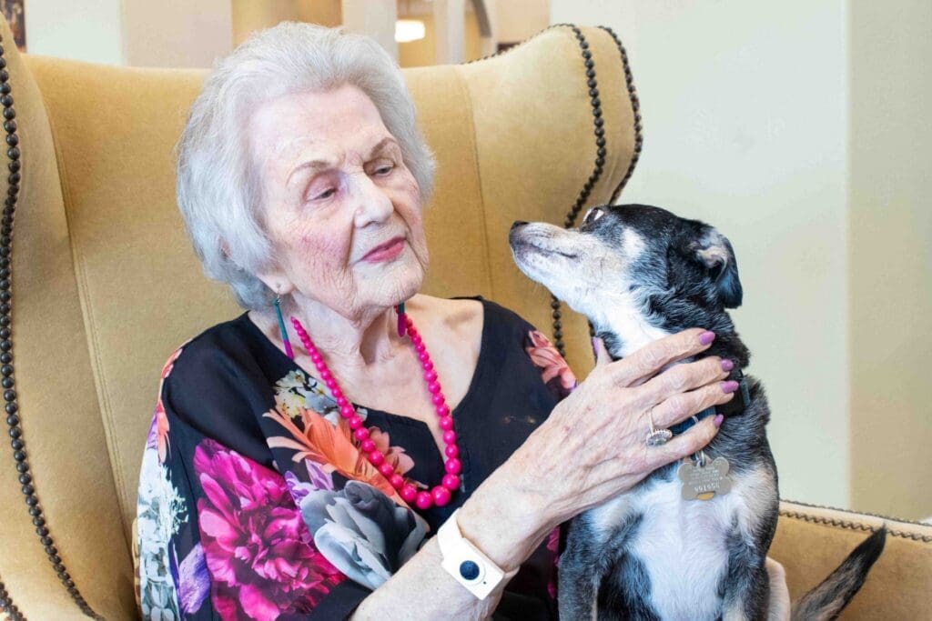 A smiling senior woman in a floral blouse lovingly holds a small dog while seated in an elegant armchair.