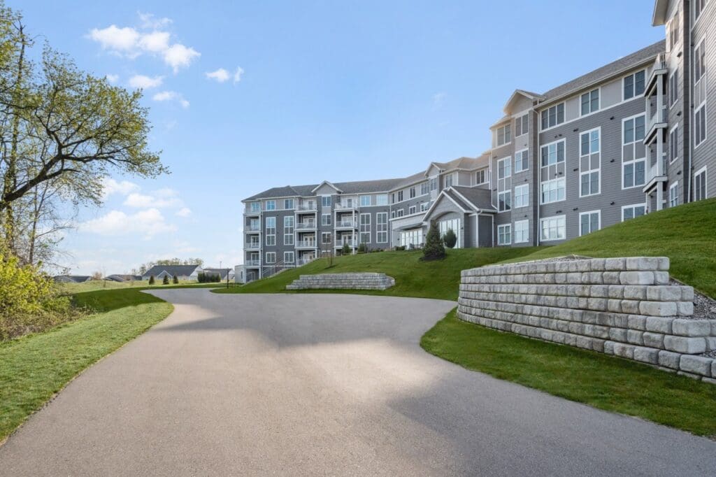 RRidge-RearExterior-01-Large - Grace Management Expansive pathway leads to a modern senior living facility, surrounded by lush greenery and welcoming outdoor spaces.