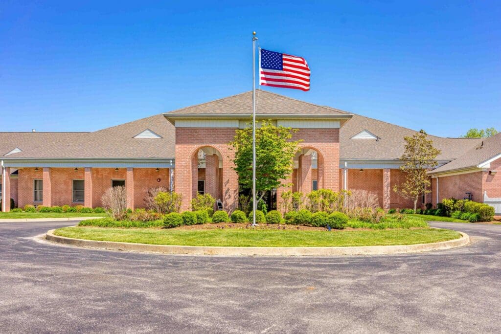 Welcoming senior living facility entrance with manicured landscaping and an American flag flying under a clear blue sky.