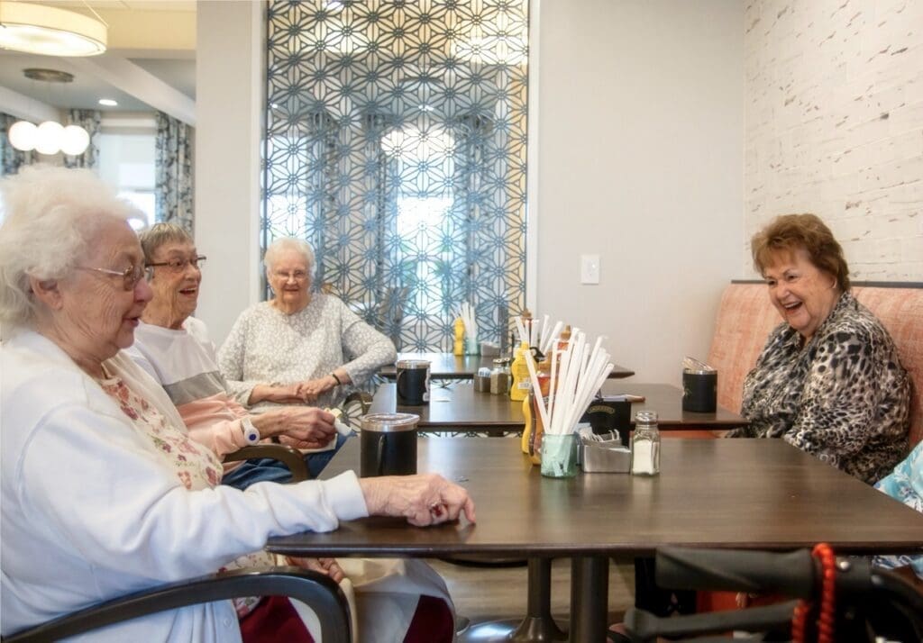 Residents-02-Large - Grace Management Four smiling senior women enjoy conversation around a table in a bright, modern café, showcasing a vibrant community atmosphere.