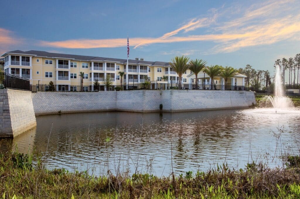 Elegant senior living community by a serene pond, featuring lush palm trees and a beautiful fountain in a tranquil setting.