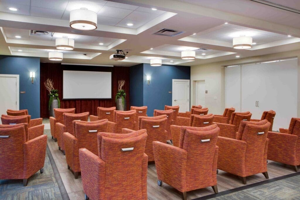 A stylish auditorium with vibrant orange chairs facing a large screen, featuring elegant lighting and potted plants.