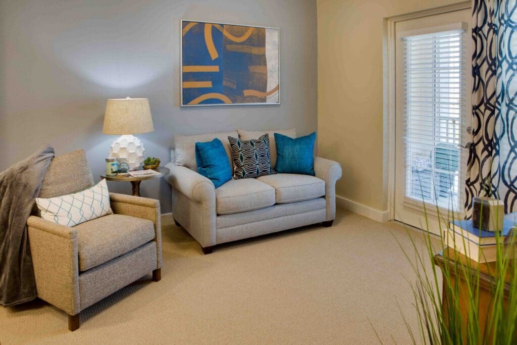 Cozy living area featuring a light gray sofa with decorative pillows, an armchair, lamp, and natural light from a patio door.