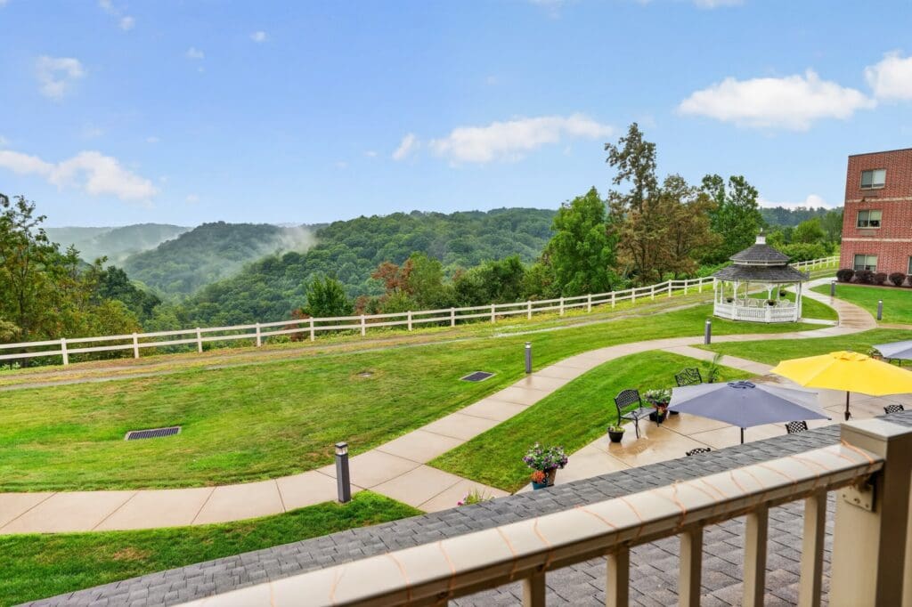 Lush green landscape with walking paths, a gazebo, and vibrant outdoor seating under umbrellas, set against serene hills.