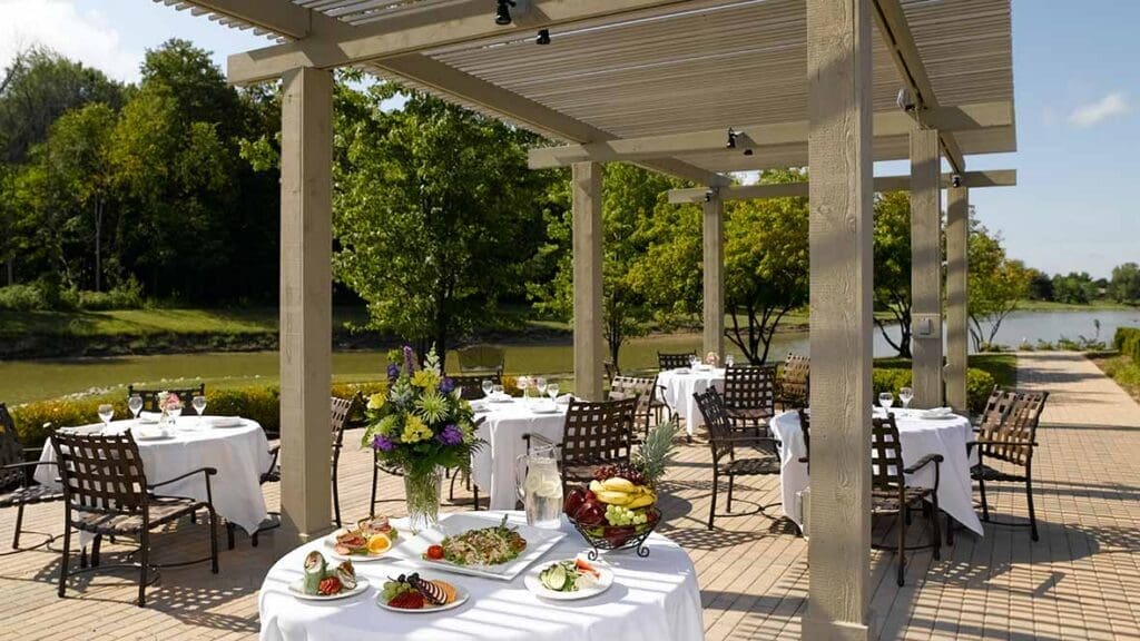 Elegant outdoor dining area featuring neatly set tables, gourmet dishes, and a tranquil lakeside view, surrounded by greenery.