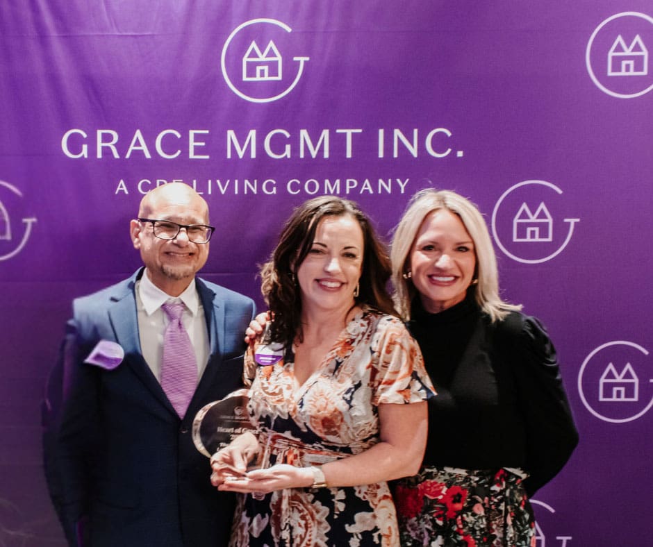 Three smiling professionals celebrate an award amidst a vibrant purple backdrop, representing Grace Management Inc.