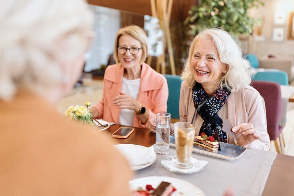 A group of lively senior women enjoy dessert and coffee together, sharing laughter in an inviting, elegant setting.
