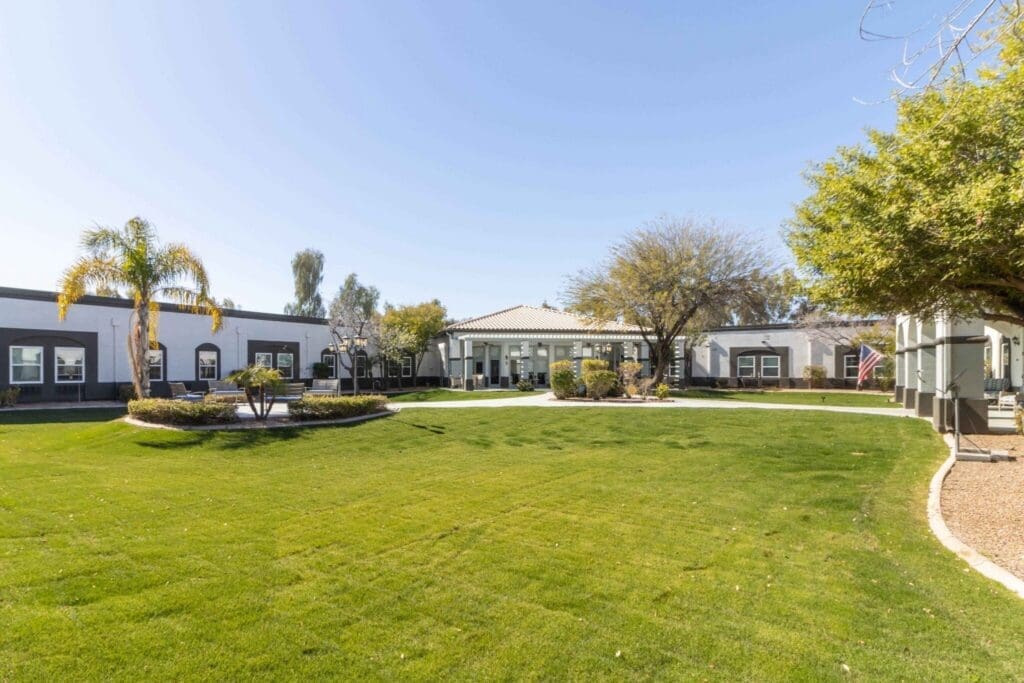 Lush green lawn with palm trees and landscaped gardens surrounding elegant senior living buildings under a clear blue sky.
