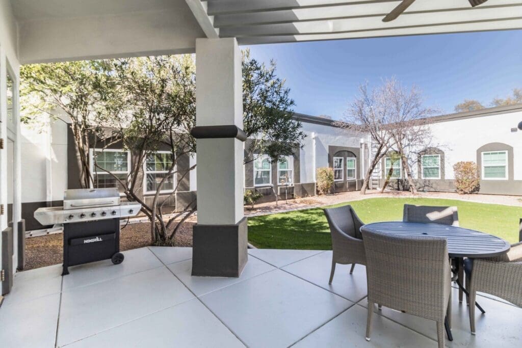 Elegant outdoor patio featuring a grill, round table, and chairs set amidst lush greenery in a serene senior living community.