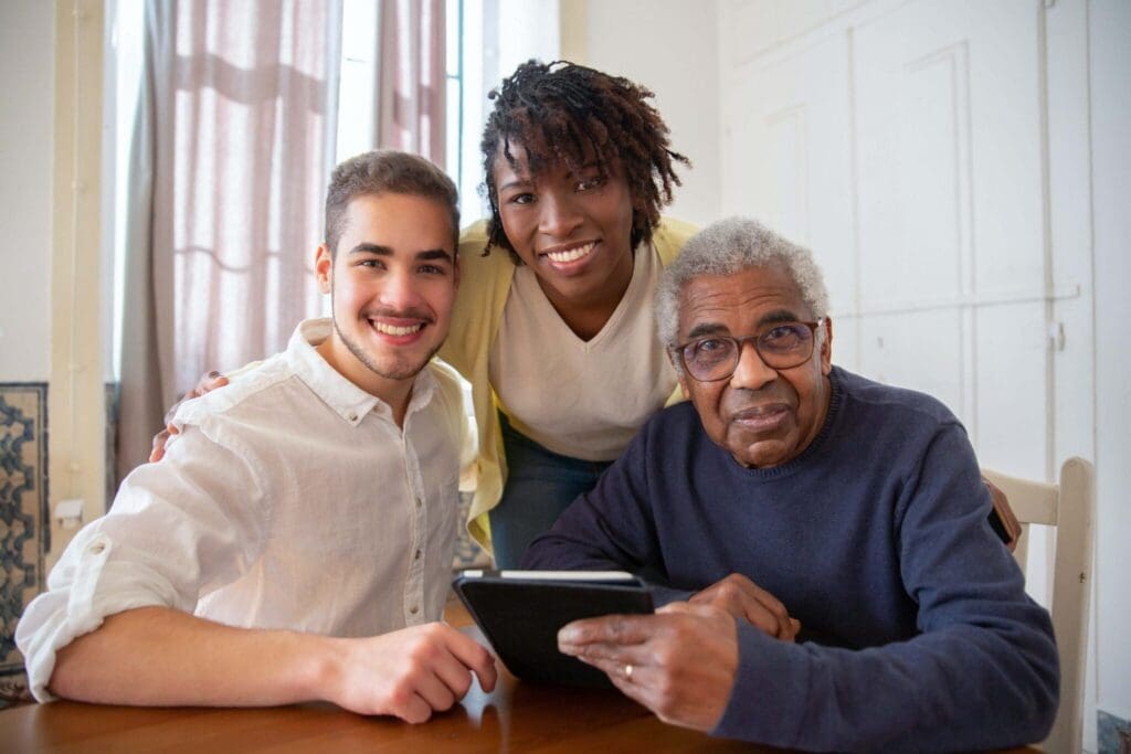 Three smiling individuals—two adults and one senior—gather around a tablet in a bright, welcoming indoor space.