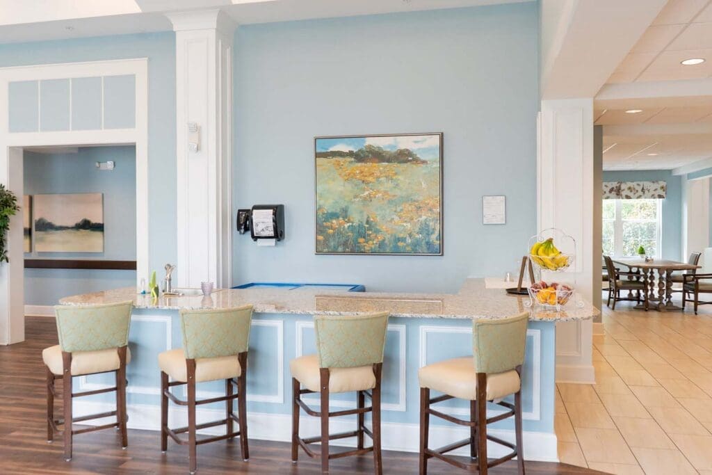the-arbors-of-gulf-breeze-32 - Grace Management Bright blue reception counter with marble top, four upholstered stools, a watercolor landscape painting on the wall, and a fruit basket on the right.