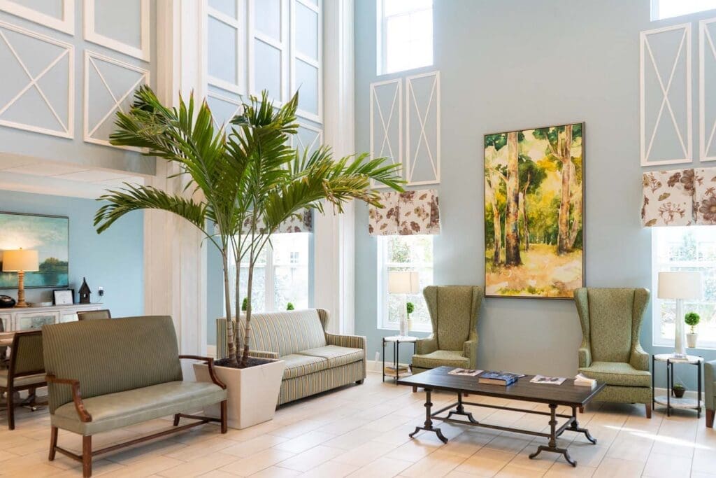 the-arbors-of-gulf-breeze-33 - Grace Management Hotel lobby seating area with light blue walls, large potted plants, and a painting above two green armchairs.