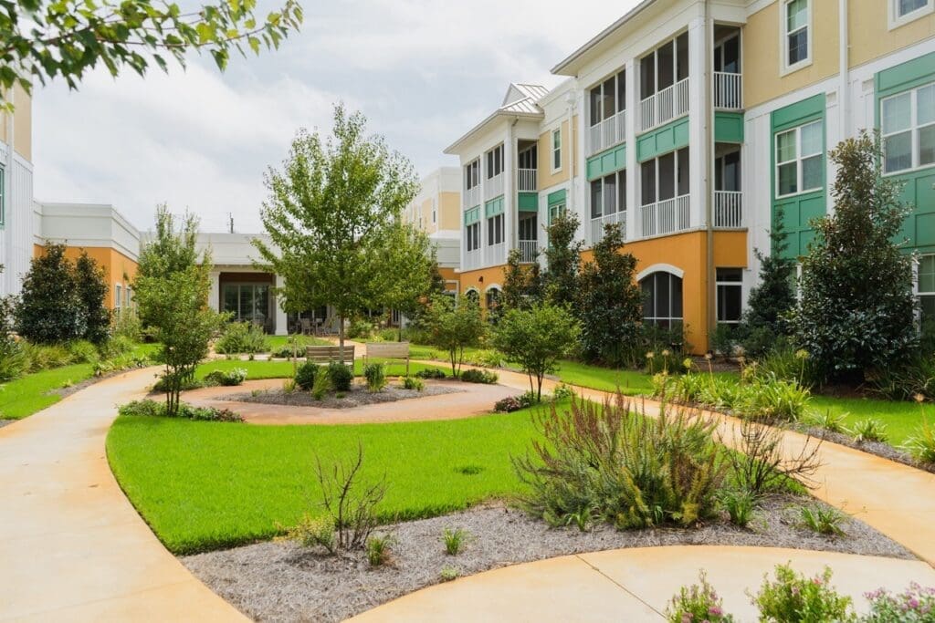 the-arbors-of-gulf-breeze-56-Large - Grace Management Residential apartment complex courtyard with green lawn, trees, and curved brick paths with benches nearby.