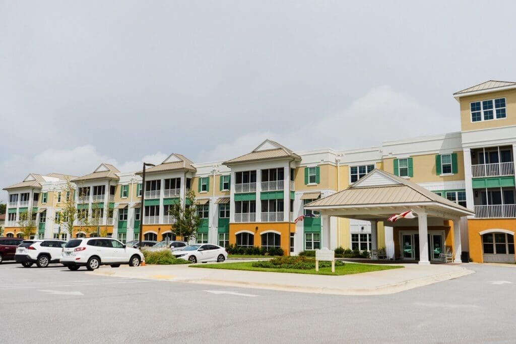 the-arbors-of-gulf-breeze-63-Large - Grace Management Colorful multi-story building with yellow and green accents and a covered entrance, resembling a hotel or residential complex; cars parked in front.