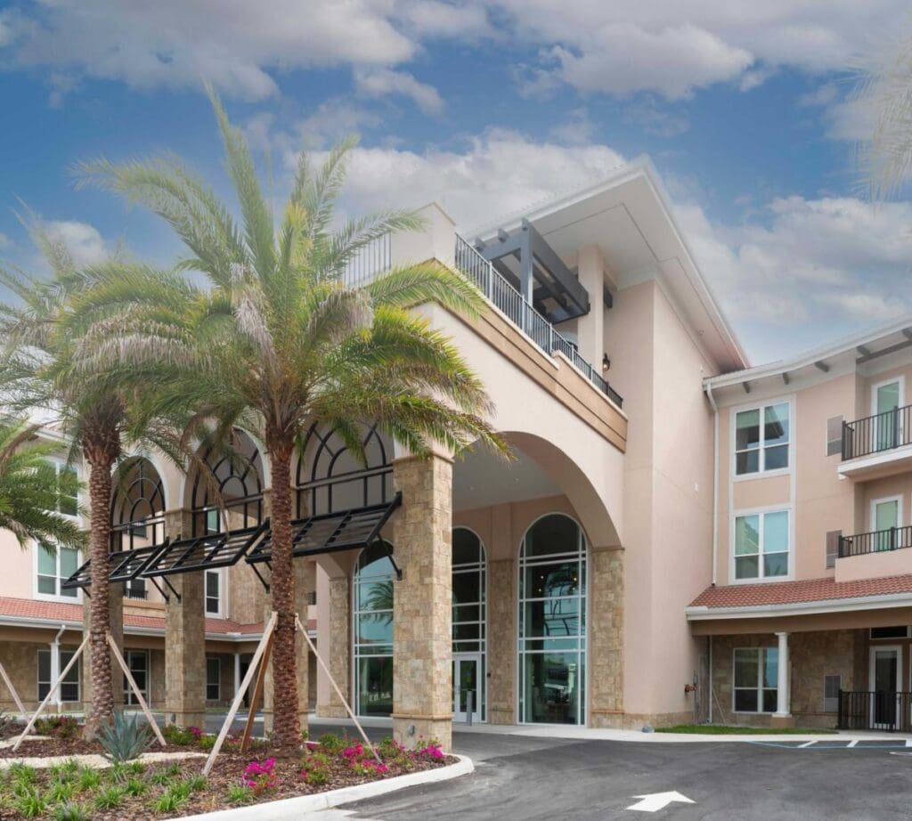 Elegant senior living community entrance framed by palm trees, featuring upscale architecture and lush landscaping.