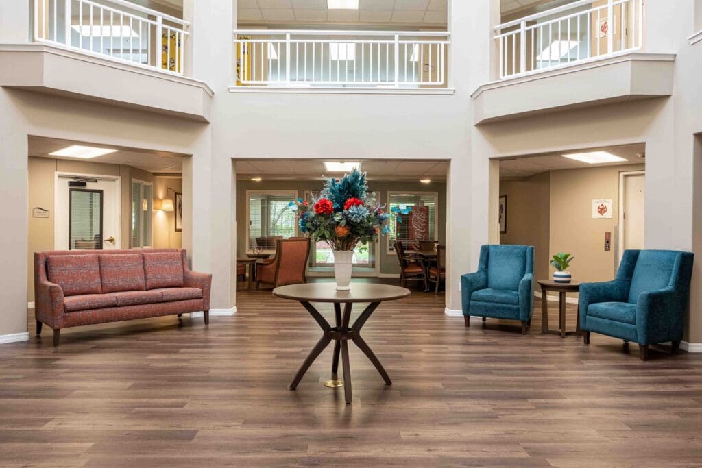 A welcoming lobby with a red couch, two blue sofa chairs, and table with a bouquet of flowers in the middle of the room at Augustine Landing, senior living community in Florida.