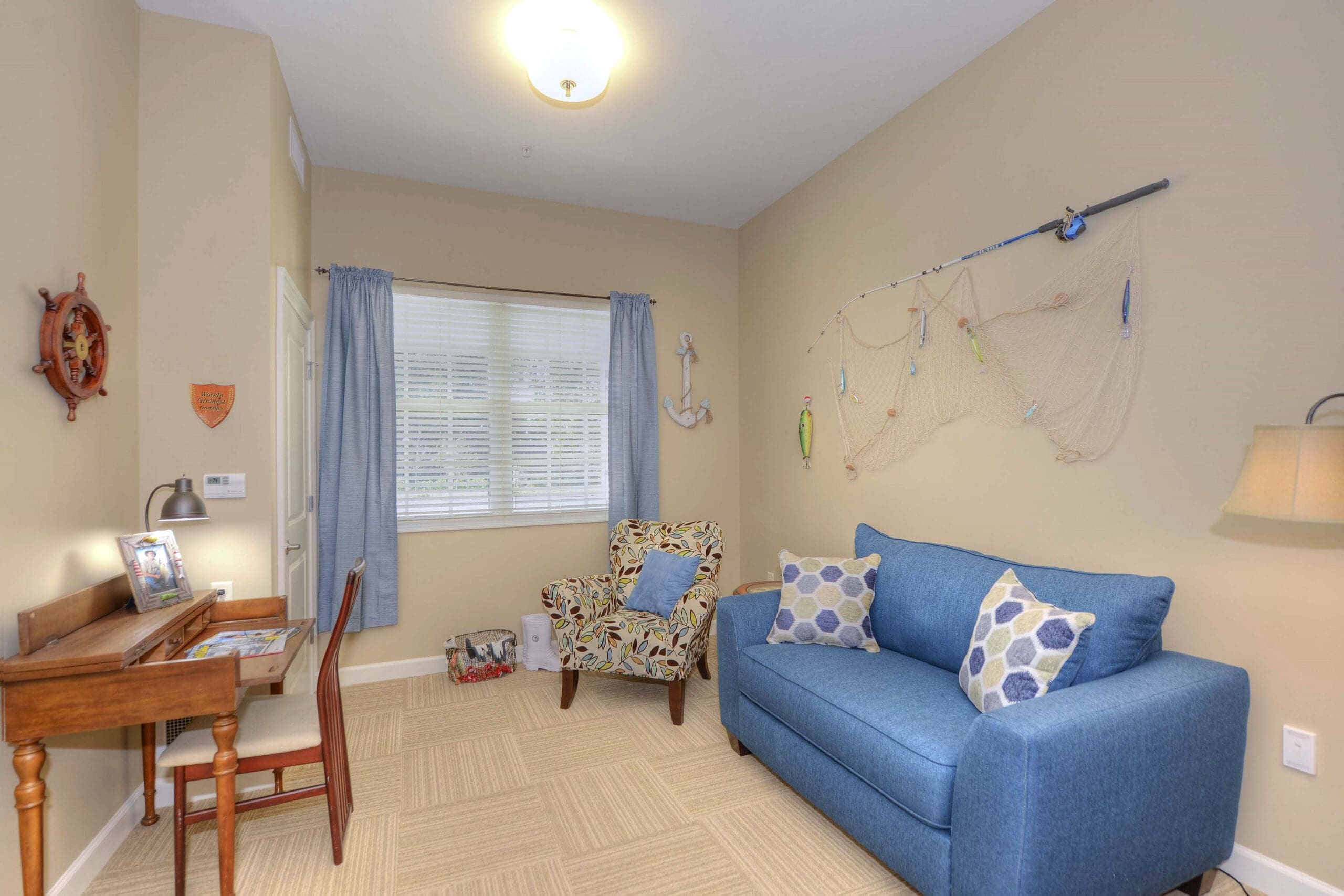 A room with a blue couch with patterned throw pillows, a patterned beige sofa chair, and a brown sitting desk with a brown chair, at Beach House, a senior living community.