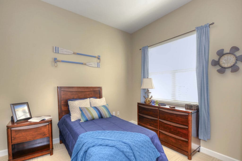 - Grace Management A bedroom with a small bed and blue sheets, throw pillows, a blue throw blanket, bedside table with a photo frame to the left of the bed, and a dresser with a lamp on it that sits underneath the window at Beach House, a senior living community.