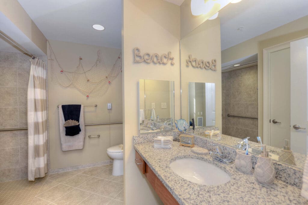 A bathroom with a marbled gray counter, mirror, sink, a walk-in shower, and a toile, Beach House senior living community.